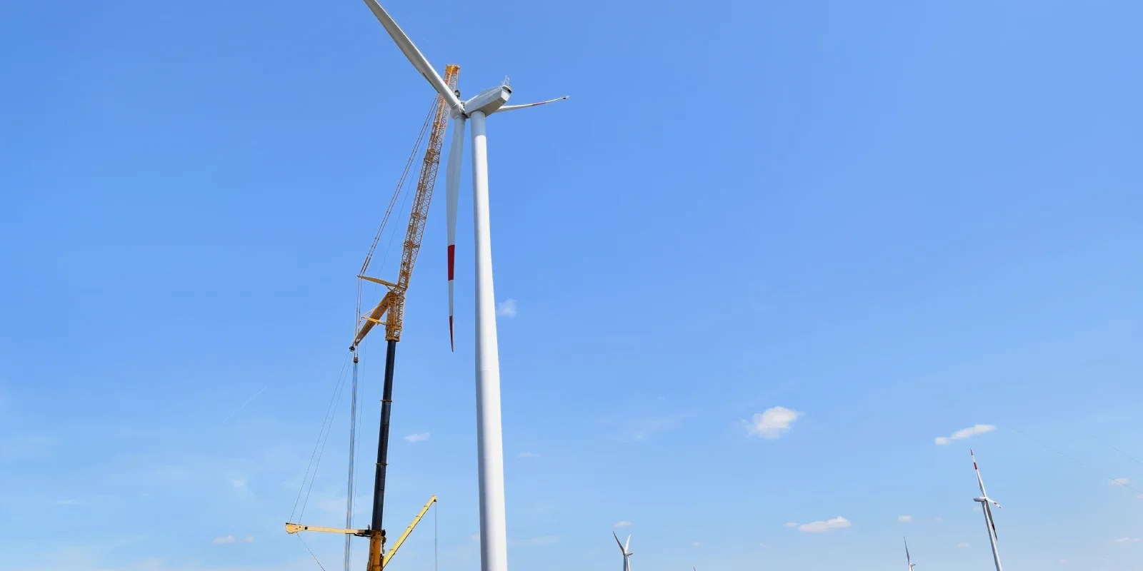 Ein Windrad mit drei weißen Rotorblättern steht vor einem klaren blauen Himmel. Die Windkraftanlage ist auf einer weiten, offenen Landschaft positioniert und die Rotorblätter sind in Bewegung zu sehen.