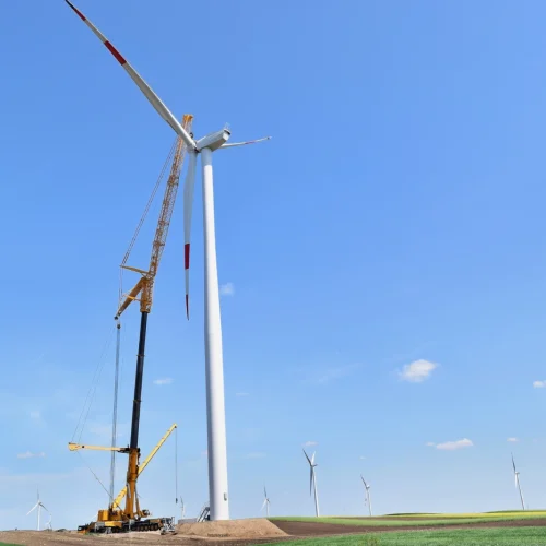 Ein Windrad mit drei weißen Rotorblättern steht vor einem klaren blauen Himmel. Die Windkraftanlage ist auf einer weiten, offenen Landschaft positioniert und die Rotorblätter sind in Bewegung zu sehen.